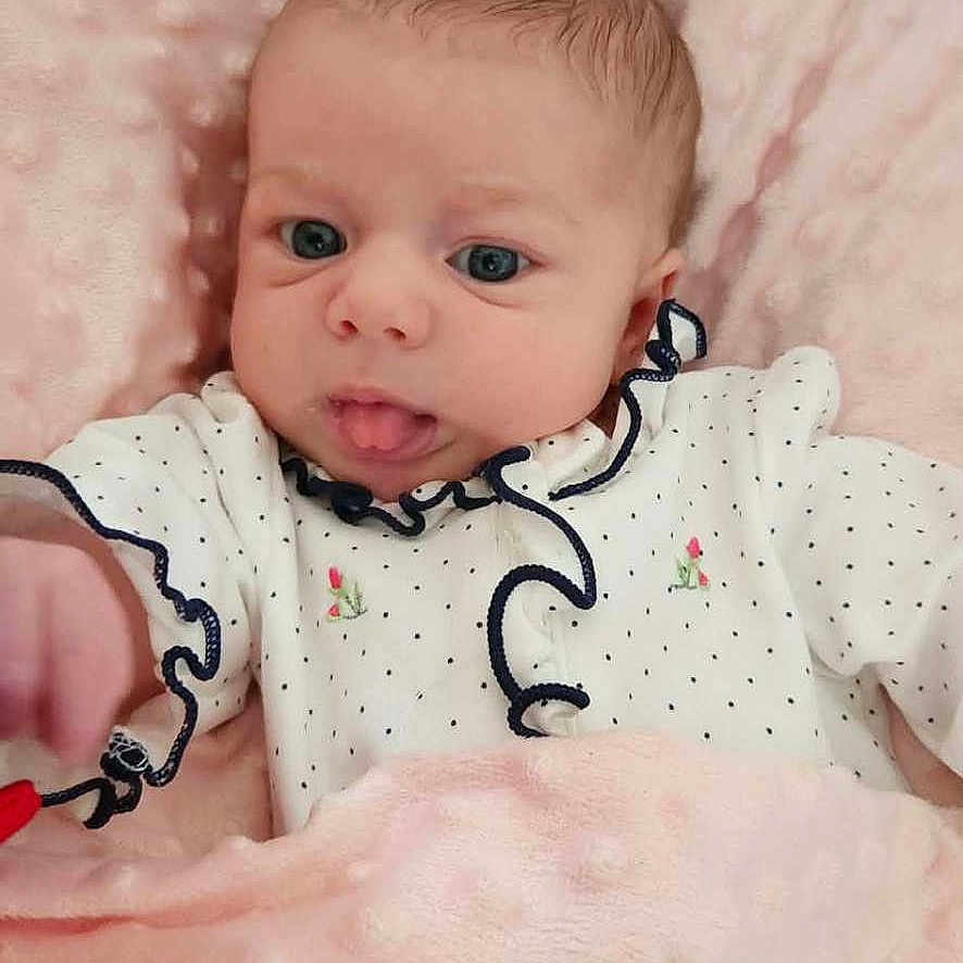 Riley joined the competition — help win amazing prizes! baby, blanket, child, clothing, comfort, cushion, cute, expression, face, floral_embroidery, home, infant, lying_down, navy_trim, newborn, pink, portrait, soft_texture, tongue_out, white_outfit