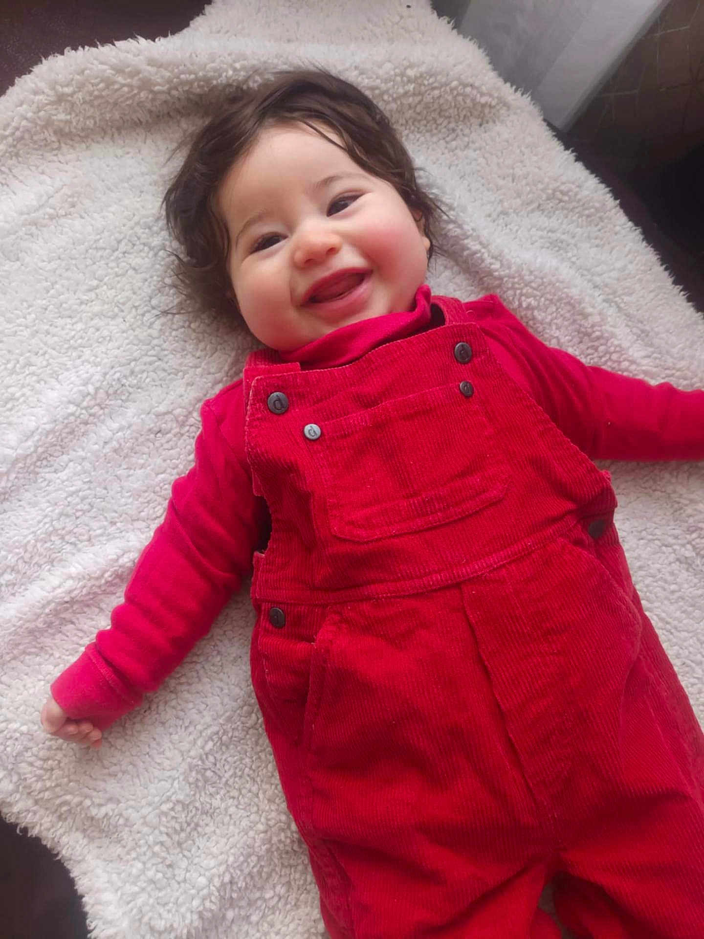 Joud a rejoint le concours — aidez-le/la à gagner de superbes lots ! baby, smiling, happy, red_overalls, curly_hair, blanket, lying_down, infant, child, cute, portrait, indoors, soft_texture, cheeks, clothing, hands, face, baby_girl, toddler, warm_colors