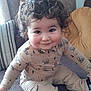 toddler, child, curly_hair, smiling, sitting, couch, pillow, beige_pants, bear_print, long_sleeve_shirt, indoor, cozy, happy, cute, face, person, clothing, home, young_child, portrait