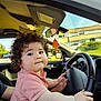 toddler, child, car, steering_wheel, curly_hair, pink_shirt, hands, adult_hand, inside_car, window, daylight, greenery, building, parked_car, seat, dashboard, rearview_mirror, cute, portrait, helping_hand