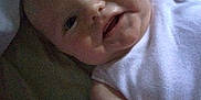 Keira Marie is registered to the contest to win money with this photo: baby, infant, face, smile, white_shirt, skin, arm, lying_down, indoors, bed, blanket, cute, child, portrait, person, young, cheeks, eyes, expression, head