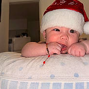 Guadalupe is registered to the contest to win money with this photo: baby, bracelet, child, closeup, curious, cute, earrings, expression, face, hand, holiday, home, indoor, infant, person, pillow, santa_hat, skin, sleeping_aid, soft