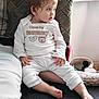 toddler, child, pajamas, white_clothing, sitting, couch, cushion, indoor, curly_hair, pony_tail, barefoot, face, looking_away, home, comfort, cute, expression, small_hands, young_child, cozy
