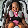 baby, smiling, car_seat, pink_clothing, pacifier, curly_hair, indoor, wooden_floor, happy, child, infant, foot, sock, seatbelt, cushion, black_hair, cute, playful, person, portrait