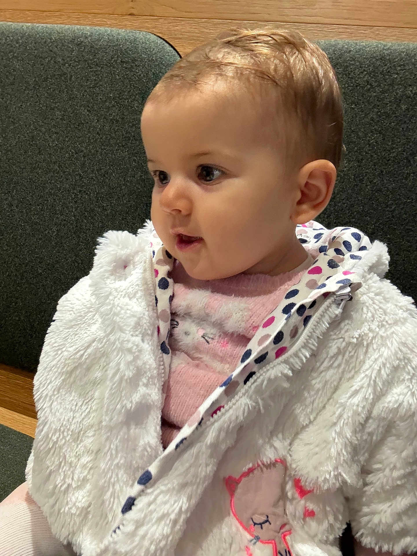 Manon participe au concours pour gagner de l'argent avec cette photo : baby, child, face, jacket, white_jacket, pink_sweater, embroidery, cat_embroidery, curly_hair, indoor, couch, green_couch, wooden_furniture, cute, warm_clothing, portrait, seated, side_view, young_child, curious