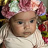 baby, flower_headband, pink_clothing, close_up, hand, face, eyes, infant, portrait, curious, skin, artificial_flowers, floral, background, person, child, cute, soft_lighting, indoors, head