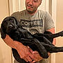 man, dog, black_dog, holding, indoor, tshirt, short_sleeve, door, content, pet, animal, smile, arms, relaxed, casual, human, friendship, companionship, cute, cozy