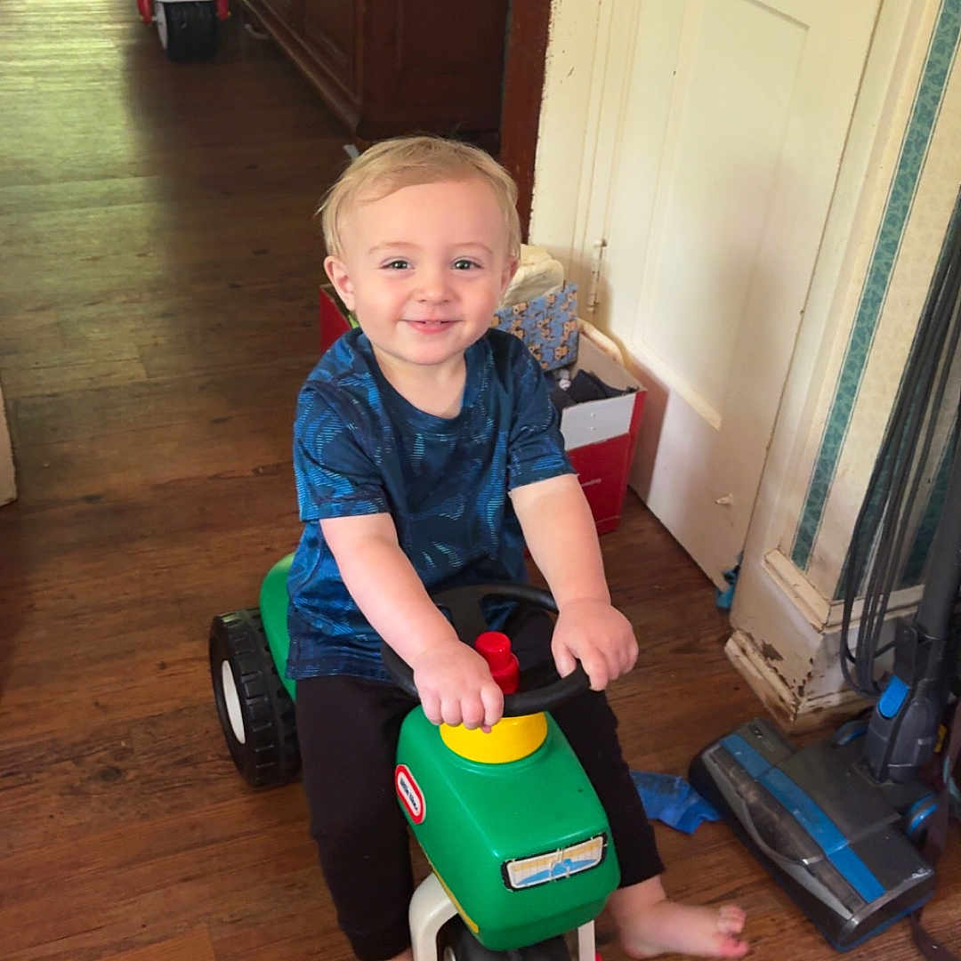 Collin Lee is registered to the contest to win money with this photo: barefoot, black_pants, blue_shirt, cabinet, child, door, flooring, happy, home, household_items, indoor, light_hair, motorcycle, playtime, smiling, toddler, toy, vacuum_cleaner, wooden_floor, young_child