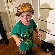 Collin Lee is registered to the contest to win money with this photo: toddler, child, indoor, hat, boots, green_tshirt, minecraft, wooden_floor, dresser, shadow, curious_expression, clothing, person, floor, door, furniture, room, standing, young_child, casual