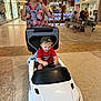 toddler, woman, toy_car, mall, shopping_center, child, indoor, people, bench, plant, floor, clothing, cap, smile, pushing, store_sign, shopping, joyful, casual, fun