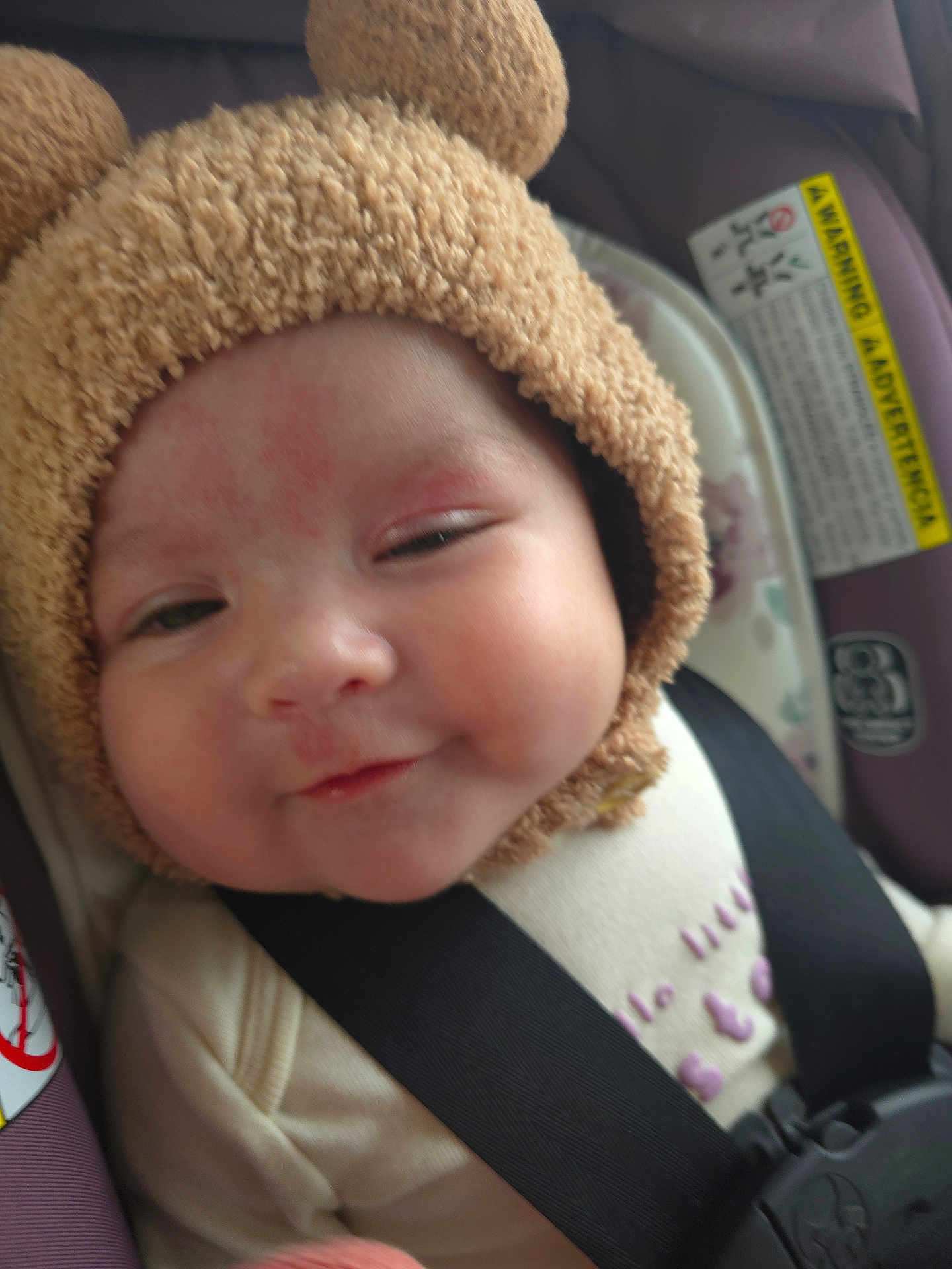 Elodie joined the competition — help win amazing prizes! baby, child, car_seat, hat, bear_ears, face, smile, infant, clothing, seatbelt, closeup, cute, redness, soft_texture, sleepy, indoors, straps, headwear, person, young