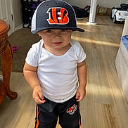 Lenox is registered to the contest to win money with this photo: toddler, child, cap, sports_cap, white_shirt, black_pants, sneakers, indoor, wooden_floor, table, fan, toy_car, curtain, carpet, person, standing, home, casual_clothing, sunlight, smile
