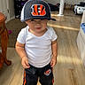 toddler, child, cap, sports_cap, white_shirt, black_pants, sneakers, indoor, wooden_floor, table, fan, toy_car, curtain, carpet, person, standing, home, casual_clothing, sunlight, smile