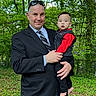 child, toddler, adult, father, parent, portrait, outdoor, forest, trees, greenery, family, holding, suit, tie, red_clothing, black_clothing, smiling, standing, photograph, smartphone_screenshot