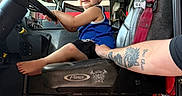 Jasper is registered to the contest to win money with this photo: child, toddler, fire_truck, vehicle_interior, steering_wheel, seated, smiling, portrait, firefighter, emergency_vehicle, red_harness, blue_shirt, black_shorts, barefoot, adult_arm, tattoo, vehicle_controls, equipment, indoors, candid_photo