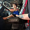 child, toddler, fire_truck, vehicle_interior, steering_wheel, seated, smiling, portrait, firefighter, emergency_vehicle, red_harness, blue_shirt, black_shorts, barefoot, adult_arm, tattoo, vehicle_controls, equipment, indoors, candid_photo