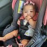 baby, child, car_seat, pink_bows, plush_toy, sucking_fingers, floral_outfit, car_interior, safety_harness, cute, portrait, infant, seatbelt, adorable, child_safety, toy, person, young_child, car_door, bracelet