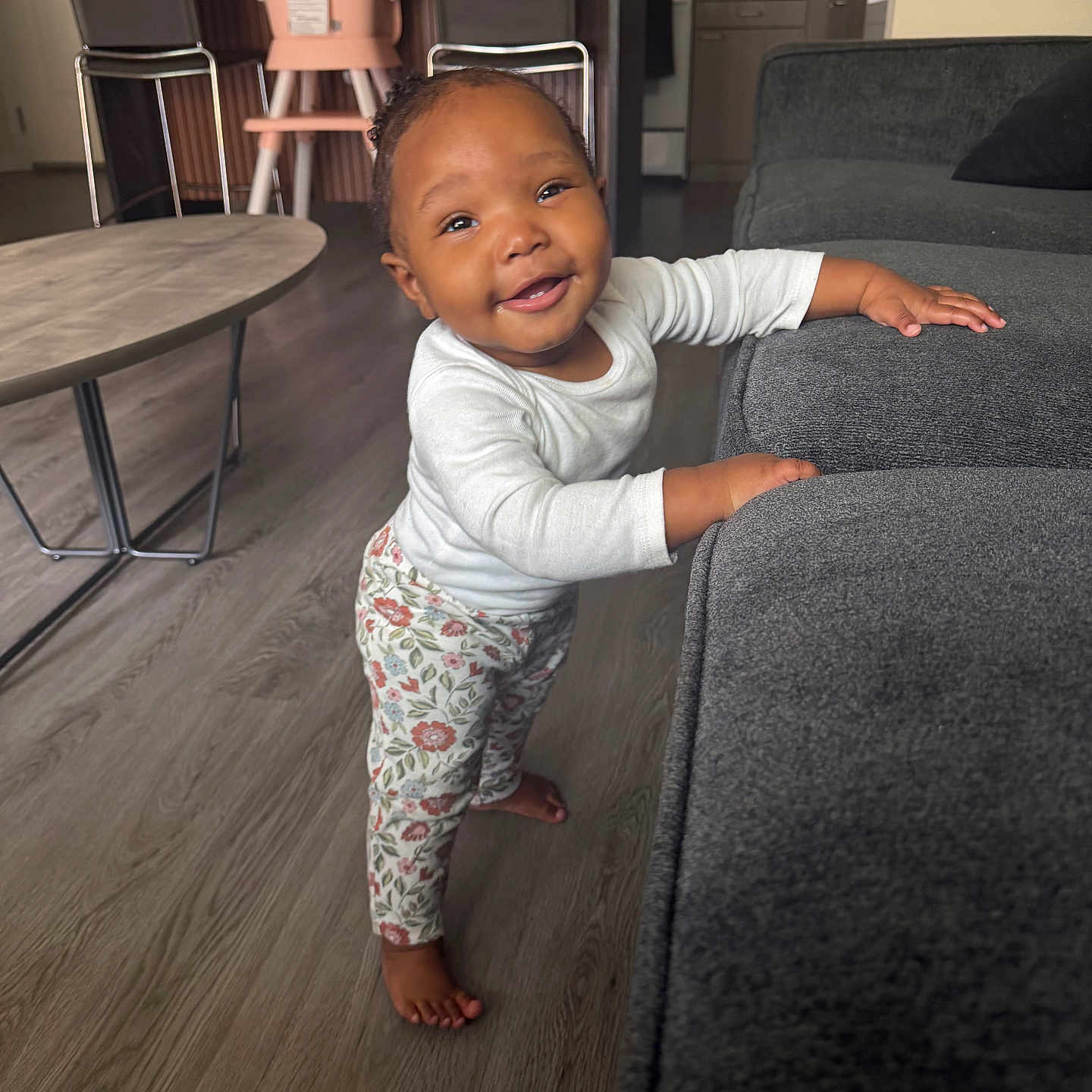 Majestic Jazelle is registered to the contest to win money with this photo: toddler, child, smiling, standing, couch, living_room, floor, table, high_chair, clothing, long_sleeve, pants, barefoot, person, home_interior, furniture, happy, portrait, baby, indoor