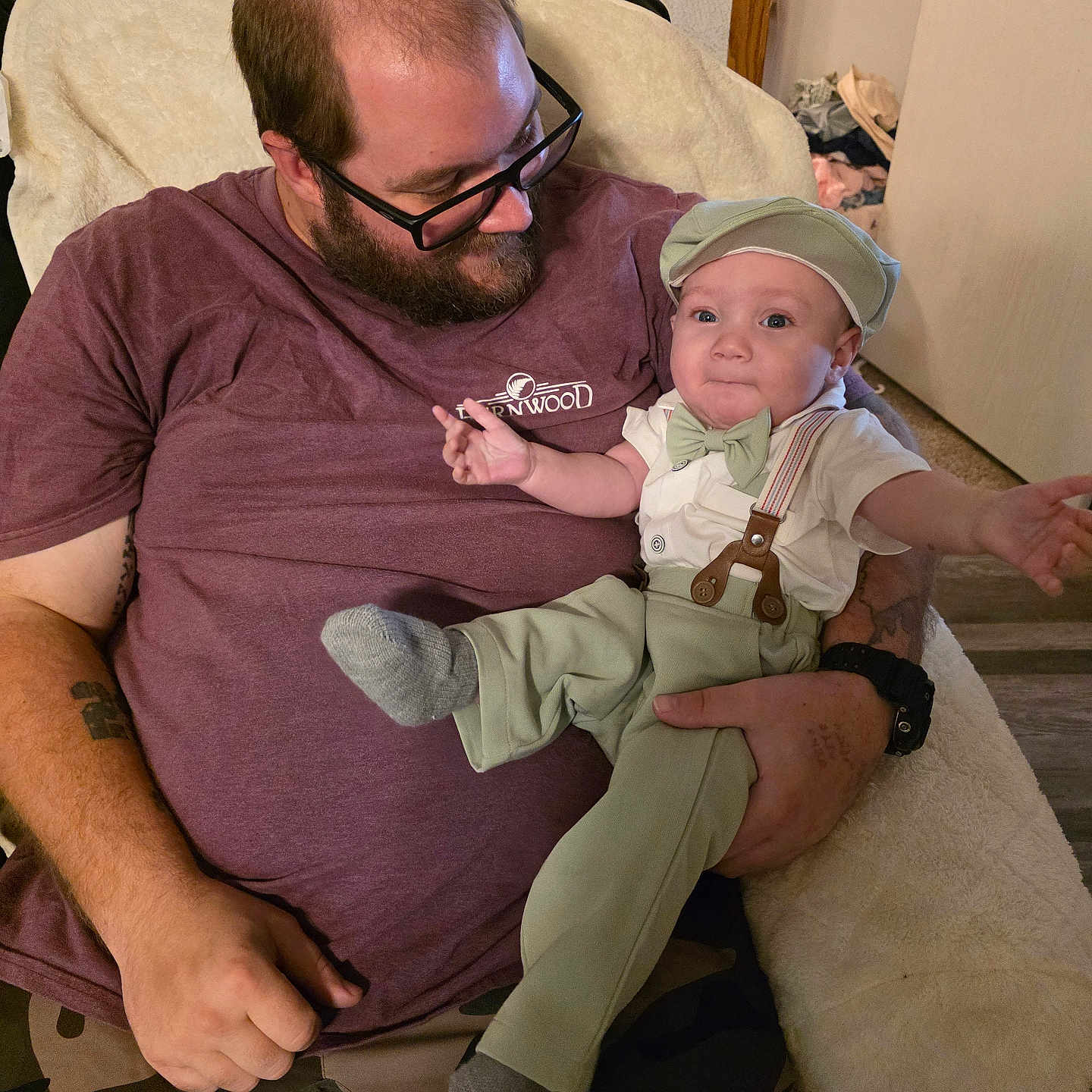 Sean joined the competition — help win amazing prizes! man, baby, child, glasses, beard, maroon_shirt, camo_pants, suspenders, bow_tie, cap, cushioned_chair, indoor, door, carpet, tattoo, watch, hand, face, smile, holding