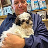 man, person, dog, puppy, small_dog, fluffy, white_fur, brown_fur, pet, pet_shop, pet_food, animal, portrait, selfie, indoors, sitting, holding, cuddling, blue_jacket, shelf