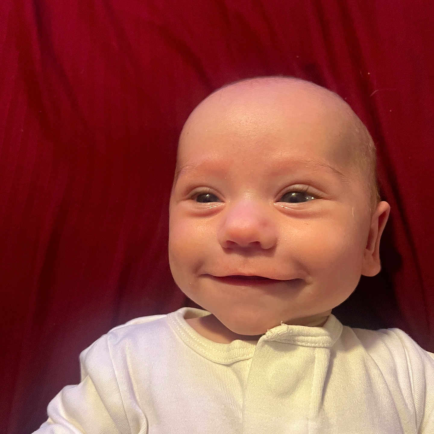 Zachary is registered to the contest to win money with this photo: baby, infant, child, portrait, smiling, face, eyes, head, white_outfit, red_background, fabric, close_up, indoor, sitting, happy, adorable, bare_head, infant_clothing, photography, close_portrait