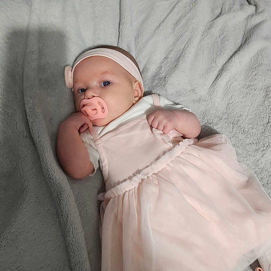 Jasmine is registered to the contest to win money with this photo: baby, infant, pacifier, pink_dress, headband, blanket, towel, bed, soft_texture, lying_down, indoors, cute, child, person, sleepwear, footwear, cozy, resting, portrait, newborn