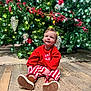 baby, child, christmas_tree, holiday, festive_clothing, red_sweatshirt, striped_pants, brown_shoes, wooden_floor, indoors, decorations, lights, pine_cones, greenery, smiling, sitting, cute, happy, portrait, seasonal