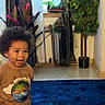 toddler, child, curly_hair, indoor, blue_rug, tshirt, plant, houseplant, window, curtain, floor, smiling, happy, standing, person, home, wall, pot, decor, casual_clothing