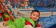 Sebastian is registered to the contest to win money with this photo: background, blanket, child, christmas, costume, cozy, cute, decor, face, festive, green_fur, grinch, holiday, indoor, lying_down, portrait, red_shirt, smiling, toddler, winter