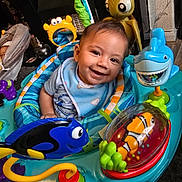 Sebastian is registered to the contest to win money with this photo: baby, smile, activity_center, toy_fish, toy_shark, toy_turtle, colorful, indoor, child, playtime, happy, infant, clothing, bib, face, seat, furniture, home, resting_baby, floor