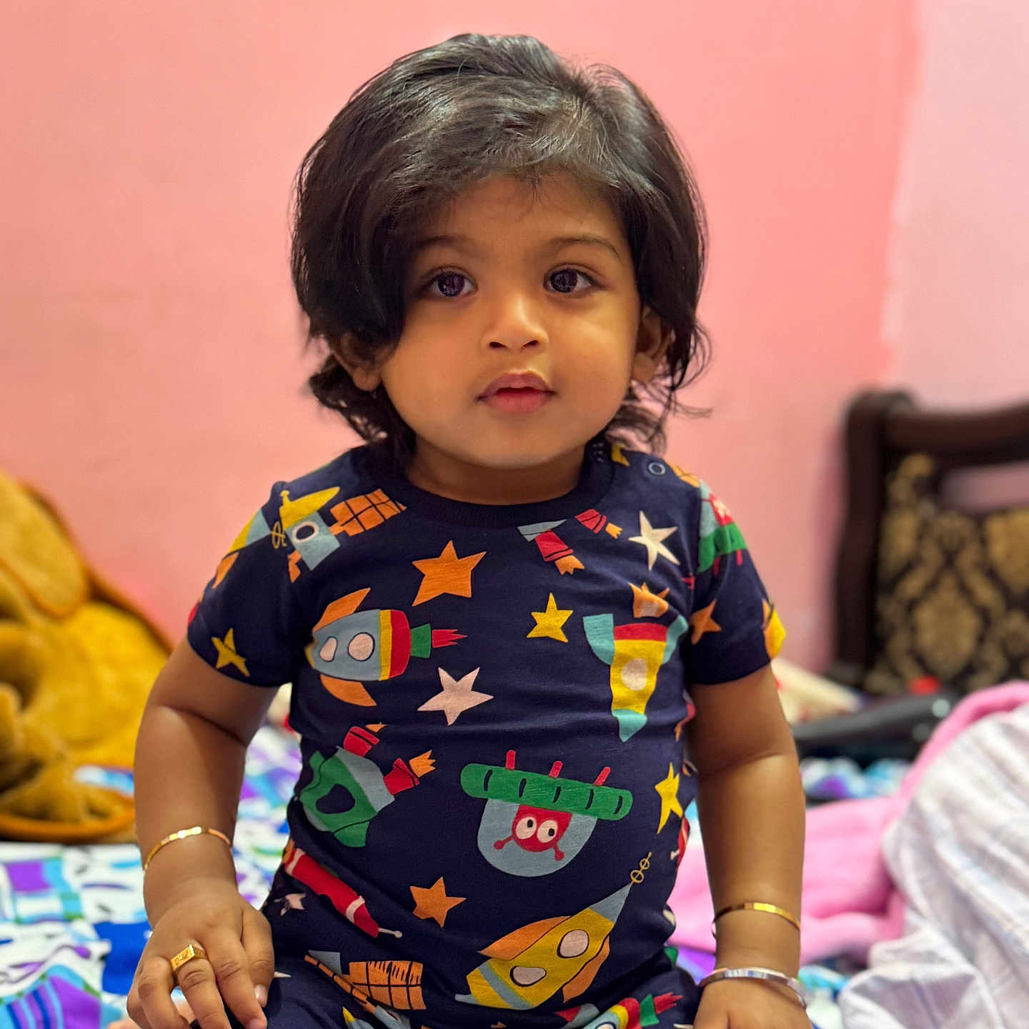 Arsh a rejoint le concours — aidez-le/la à gagner de superbes lots ! bed, blanket, bracelets, child, colorful_clothing, curly_hair, cute, furniture, indoor, looking_at_camera, pink_wall, portrait, rings, rocket_pattern, room, short_sleeves, sitting, stars, toddler, young_child