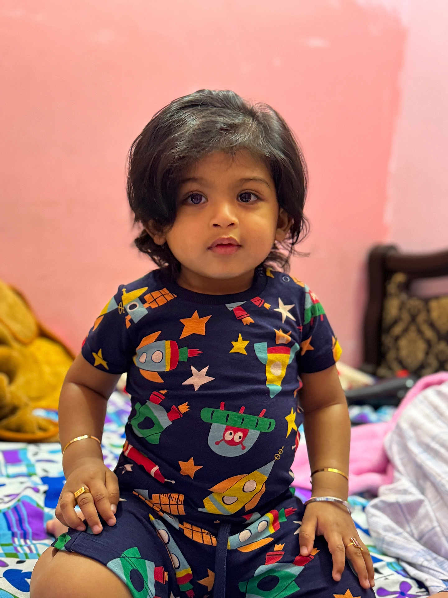 Arsh a rejoint le concours — aidez-le/la à gagner de superbes lots ! bed, blanket, bracelets, child, colorful_clothing, curly_hair, cute, furniture, indoor, looking_at_camera, pink_wall, portrait, rings, rocket_pattern, room, short_sleeves, sitting, stars, toddler, young_child