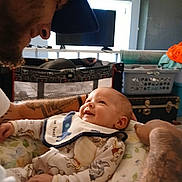 Ayden is registered to the contest to win money with this photo: baby, adult, tattoo, smile, bib, playpen, television, furniture, room, clothing, happy, face, person, interaction, home, infant, closeup, portrait, indoor, parent