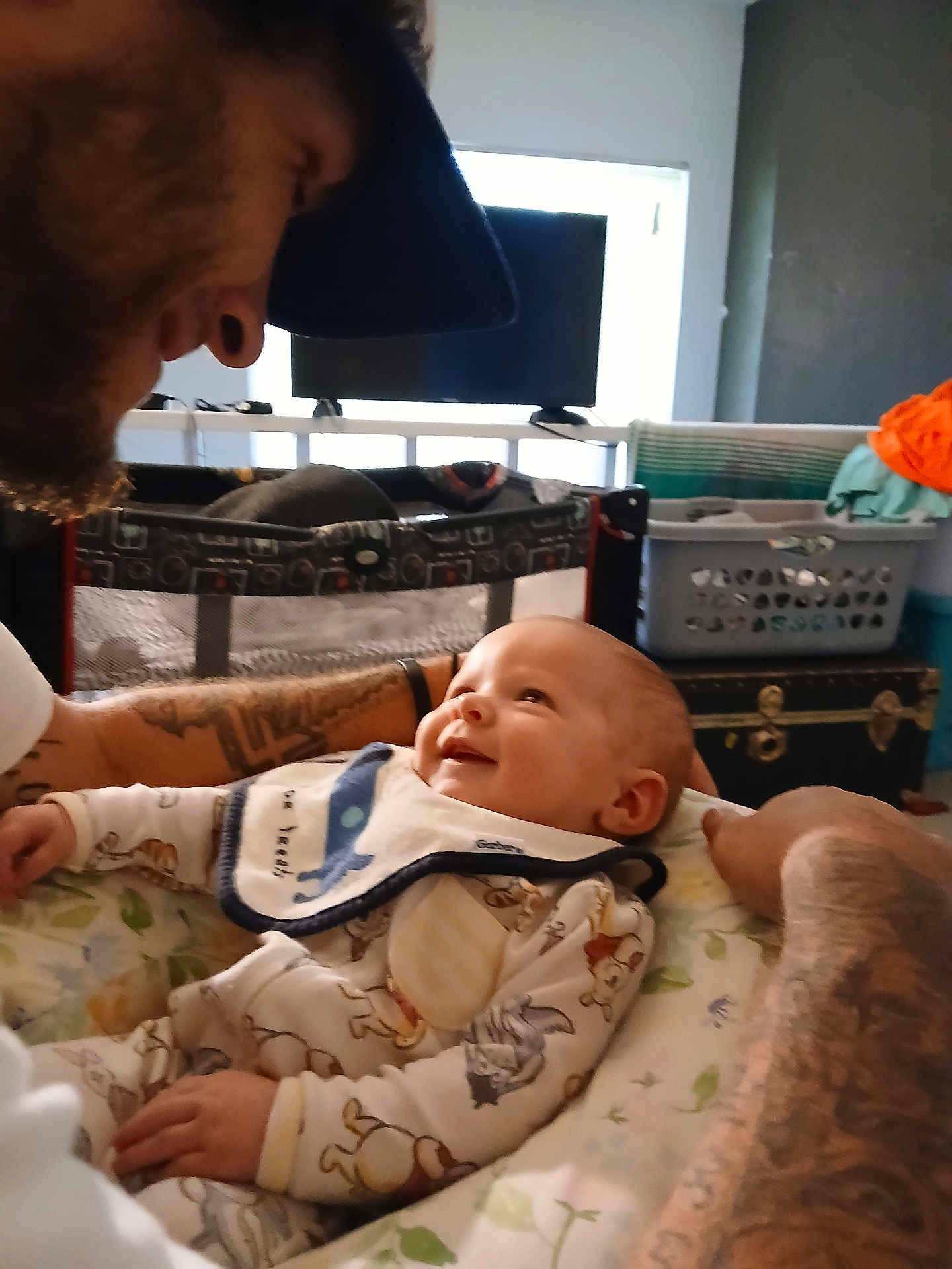Ayden is registered to the contest to win money with this photo: baby, adult, tattoo, smile, bib, playpen, television, furniture, room, clothing, happy, face, person, interaction, home, infant, closeup, portrait, indoor, parent