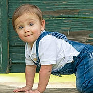 Cruz is registered to the contest to win money with this photo: toddler, child, crawling, denim_overalls, white_shirt, outdoor, green_background, wooden_wall, cute, curious, baby, person, portrait, young_child, casual_clothing, hands, knees, face, expression, little_kid