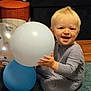Richie is registered to the contest to win money with this photo: toddler, child, balloon, blue_balloon, white_balloon, carpet, smiling, happy, indoors, wrapped_gifts, floor, light_blonde_hair, baby_clothes, sitting, playful, person, home, joyful, toy, celebration
