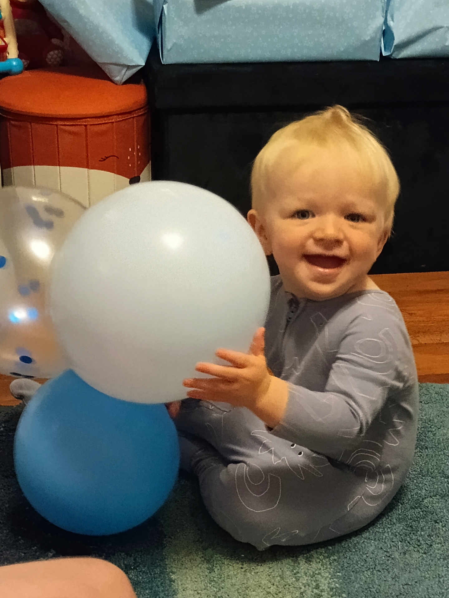 Richie is registered to the contest to win money with this photo: toddler, child, balloon, blue_balloon, white_balloon, carpet, smiling, happy, indoors, wrapped_gifts, floor, light_blonde_hair, baby_clothes, sitting, playful, person, home, joyful, toy, celebration