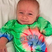 Tucker joined the competition — help win amazing prizes! baby, infant, smile, tie_dye_shirt, colorful_clothing, text_on_clothing, pillow, white_background, hand, face, head, person, cute, happy, portrait, indoors, child, newborn, young_child, casual_clothing