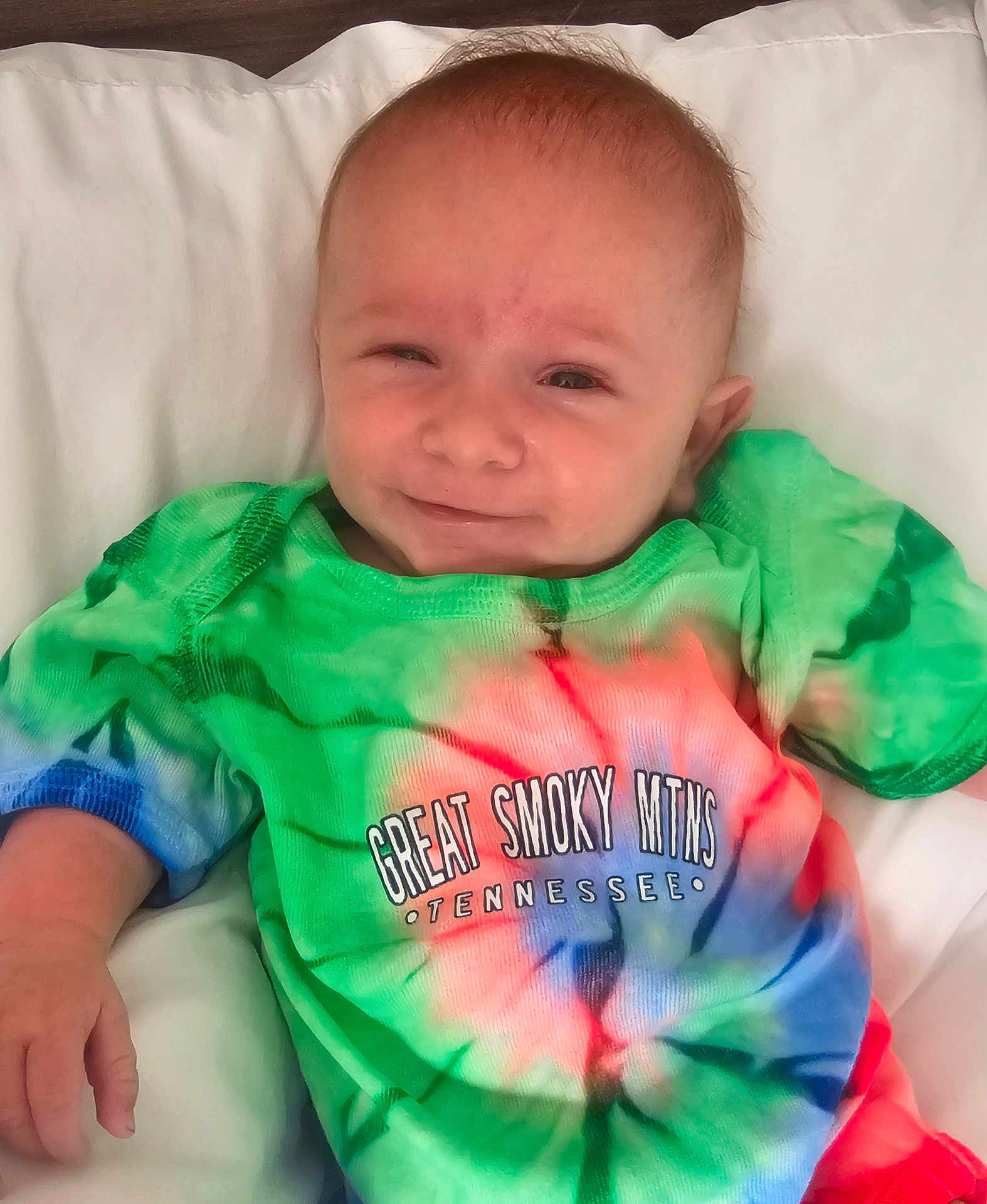 Tucker joined the competition — help win amazing prizes! baby, infant, smile, tie_dye_shirt, colorful_clothing, text_on_clothing, pillow, white_background, hand, face, head, person, cute, happy, portrait, indoors, child, newborn, young_child, casual_clothing
