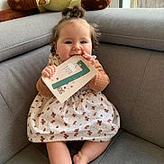 Aliénor participe au concours pour gagner de l'argent avec cette photo : baby, barefoot, child, couch, curly_hair, dress, face, foot, hand, happy, home, indoor, milestone_card, patterned_clothing, plush_toy, seated, skin, smiling, teddy_bear, window