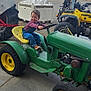 atv, boy, child, clouds, concrete_floor, farm_equipment, field, green_tractor, outdoor, playful, quad_bike, rural, seat, sky, smiling, steering_wheel, tire, tractor, wheel, workshop