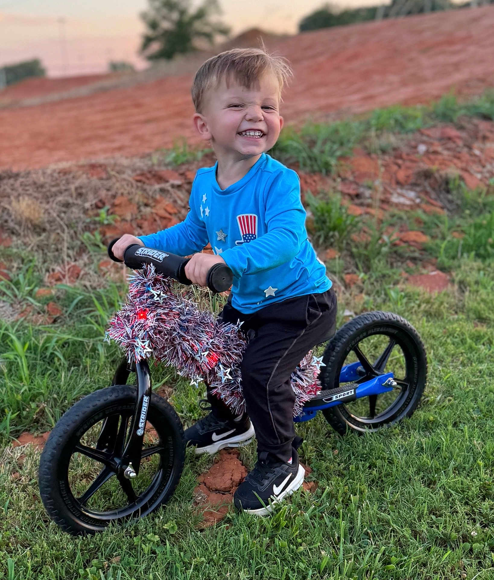 Houston Lane joined the competition — help win amazing prizes! child, smiling, balance_bike, tinsel, stars, outdoor, grass, dirt, blue_shirt, black_pants, black_shoes, nike, happy, young_boy, playing, toy, sunset, nature, fun, portrait