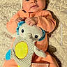baby, bow_headband, child, close_up, cushion, cute, elephant_toy, face, feet, hands, indoor, infant, lying_down, orange_outfit, patterned_fabric, photography, plush_toy, portrait, smiling, toy