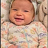 baby, infant, child, smiling, portrait, floral_outfit, headband, bow, onesie, blanket, plush_toy, bunny, cuddly, happy, indoors, close_up, sitting, face, hands, cozy