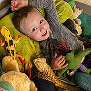 Elijah is registered to the contest to win money with this photo: child, smiling, stuffed_animal, plush_toy, bearded_dragon, reptile, toy_lizard, bedroom, cushion, green_toy, gray_shirt, blue_eyes, happy, indoor, soft_toy, boy, playful, colorful, hand, lying_down