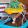 toddler, child, baby_walker, green_shirt, yellow_tray, blue_base, wooden_floor, smiling, indoor, toy, footwear, shorts, person, young_child, happy, playtime, walker, furniture, home, cute