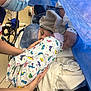 Brianna is registered to the contest to win money with this photo: newborn, baby, mother, hospital, medical_staff, mask, blanket, dinosaur_pattern, bed, gown, hand, face, hat, support, care, person, medical_equipment, blue_drape, tenderness, delivery_room