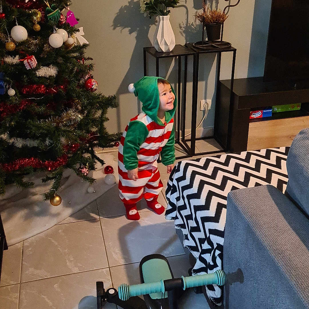 Cally participe au concours pour gagner de l'argent avec cette photo : blanket, child, christmas_tree, couch, decor, festive_clothing, floor_tiles, flowers, furniture, happy, holiday, hooded_onesie, indoor, living_room, present, shadow, smile, toddler, toy_bike, vase
