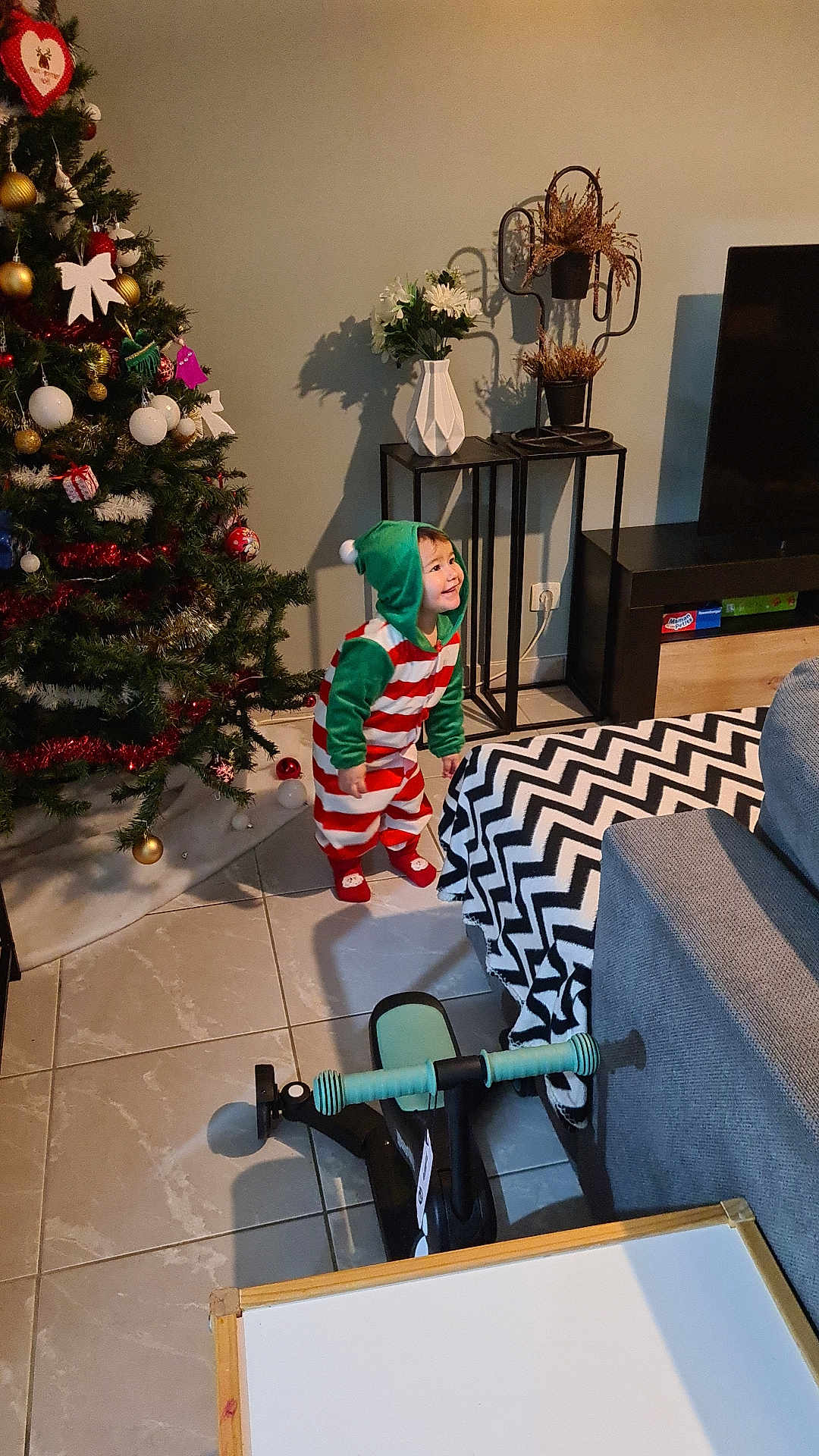 Cally participe au concours pour gagner de l'argent avec cette photo : toddler, child, christmas_tree, holiday, festive_clothing, living_room, toy_bike, blanket, couch, floor_tiles, vase, flowers, decor, smile, hooded_onesie, indoor, furniture, shadow, present, happy