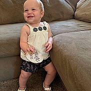 Zaria is registered to the contest to win money with this photo: toddler, child, baby, smiling, standing, living_room, couch, carpet, white_top, shorts, sandals, wooden_sign, nine_months, happy, indoor, person, portrait, cute, clothing, home